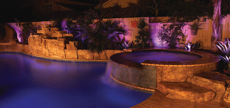 Color-lit backyard pool and spa with purple and amber landscape lighting on rock wall and plants